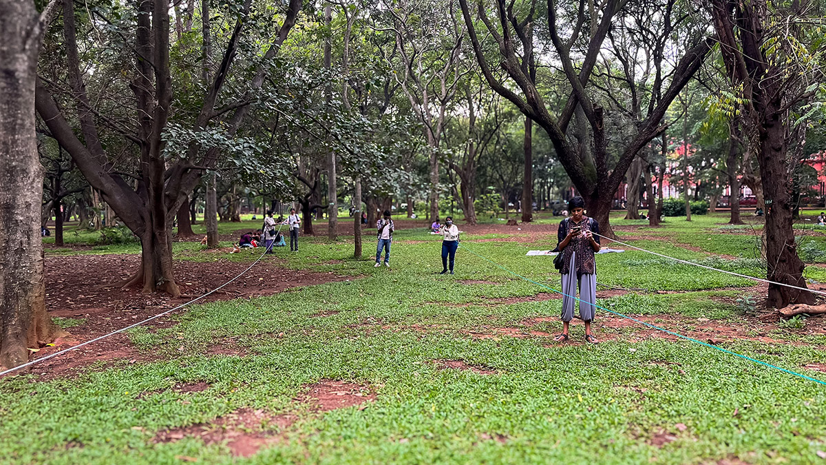 A group of people at Cubbon Park participate in a workshop activity in which they measure the dimensions of a hotspot. They are holding ropes stretched between trees. The park features tall trees, dense foliage, and a carpet of grass.