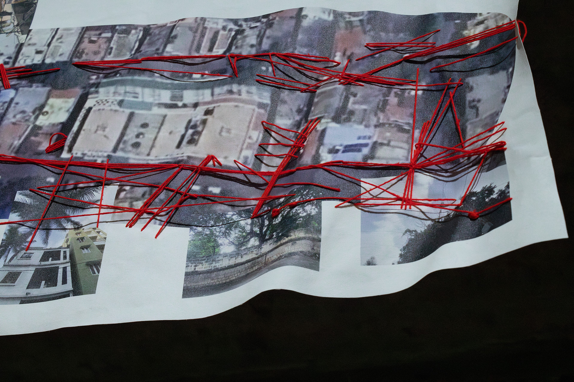 A closeup shows parts of the work Yarn and Wires. The work consists of a canvas with a print of two streets captured through the Satellite view mode of Google Maps with all internet and electricity cables visible in these streets stitched atop. The Map view is overlayed with photographs of what the streets look like, showing for example a house, a wall with a tree behind, and a tree photographed from below. The red yarn is stitched across the fabric, coming in and out at different positions, some of them near roofs, trees and electricity poles. The work was photographed while it was moved by the wind, and the lower edge is captured while it is wavy.