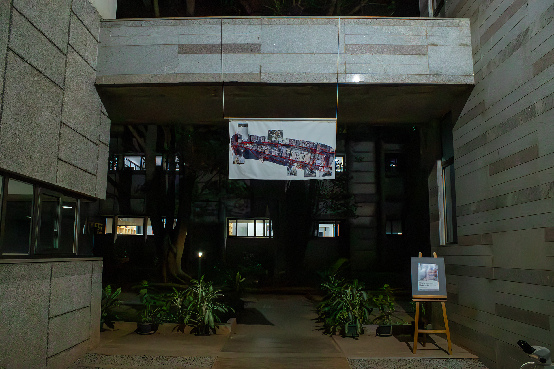 A photo taken during the opening night shows the work Yarn and Wires. The banner is about 1.5m times 0.8 m in dimensions, and has the screenshot of two streets as captured on Google Maps Satellite mode printed across. With red yarn, all cables in the streets are stitched atop. The work is hanging from a concrete bridge in the Southern Laboratories building at the National Center for Biological Sciences Bangalore. Below, plants in pots are positioned around a pathway that one can follow to move underneath and beyond the banner. The darkness in the background is interrupted by the lights of windows.